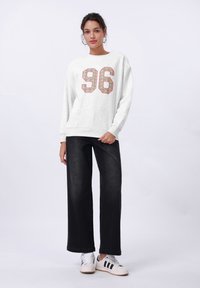 Felpa bianca con una grande stampa luccicante "96", abbinata a jeans neri a gamba larga e sneakers bianche con strisce nere.