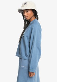Felpa oversized azzurro chiaro con maniche lunghe, orlo a costine e logo ricamato. Abbinata a un cappello a secchiello texturizzato color crema.