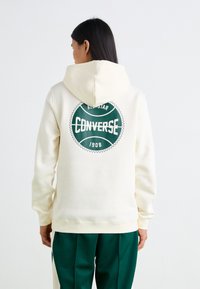 Sudadera crema con un gran logo circular verde de Converse en la espalda. Cuenta con capucha ajustable y puños acanalados, diseñada para un ajuste relajado.