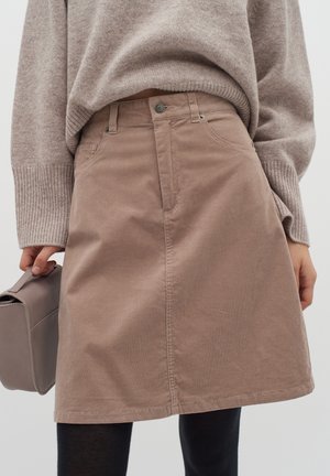 Femme portant une jupe beige en velours côtelé de forme trapèze, un pull beige ample, des collants noirs et tenant un sac taupe à côté d'elle.