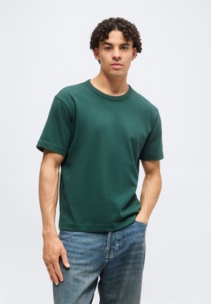 Ung mand med krøllet hår iført en mørkegrøn t-shirt med korte ærmer og blå jeans, stående mod en ensfarvet lys baggrund.
