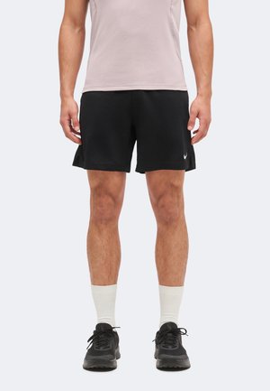 Männlich trägt schwarze Nike-Sportshorts, weiße Kniestrümpfe, schwarze Sneakers und ein hellrosa ärmelloses Oberteil, steht mit entspannten Armen.