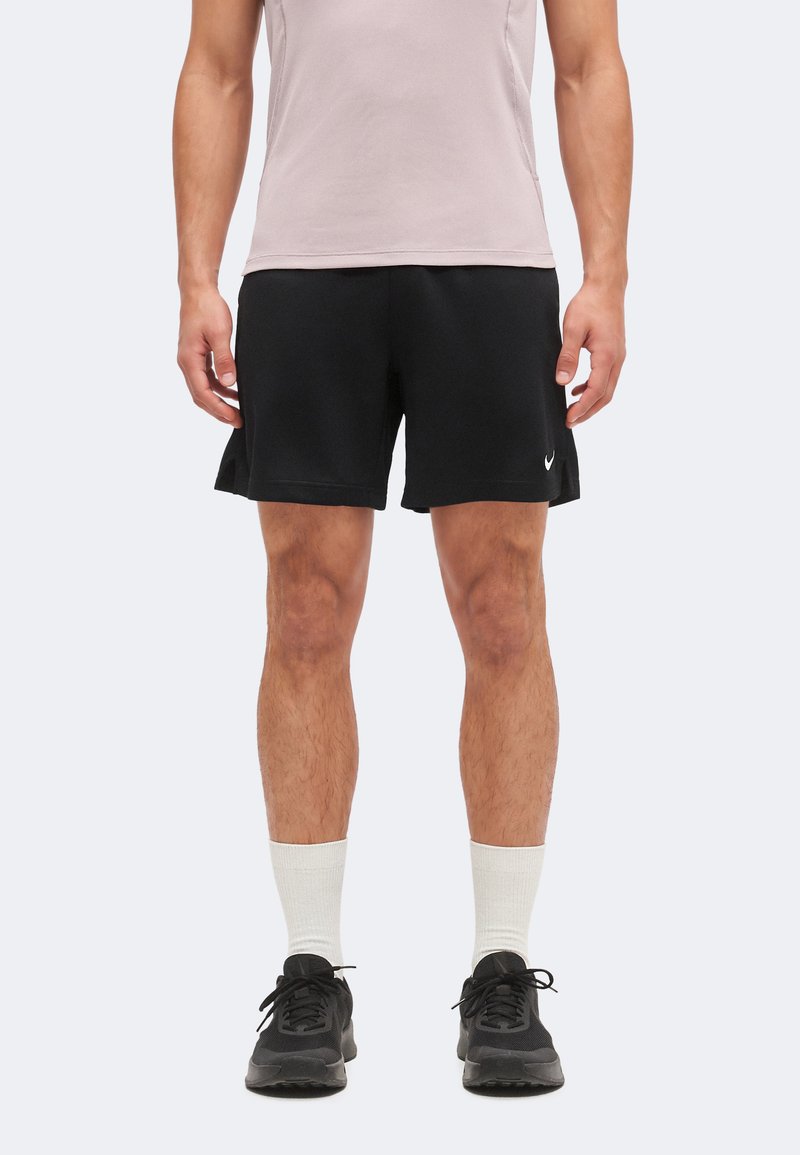 Άνδρας που φοράει μαύρο αθλητικό σορτσάκι Nike, λευκές κάλτσες μεσαίου μήκους, μαύρα αθλητικά παπούτσια και αμάνικο ροζ ανοιχτό τοπ, στέκεται με χαλαρά χέρια.