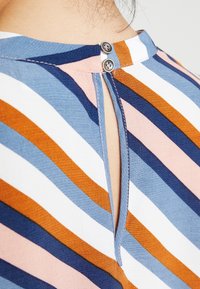 Gestreifte Bluse in Blau, Orange, Pink und Weiß. Rückenschluss mit zwei metallischen Knöpfen und einem kleinen Schlüssellochdetail.