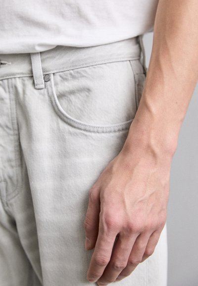 Pantaloni in denim grigio chiaro con passanti per cintura, chiusura con bottone e dettagli sui taschini discretamente lavorati. Il tessuto ha un aspetto liscio e leggermente strutturato.