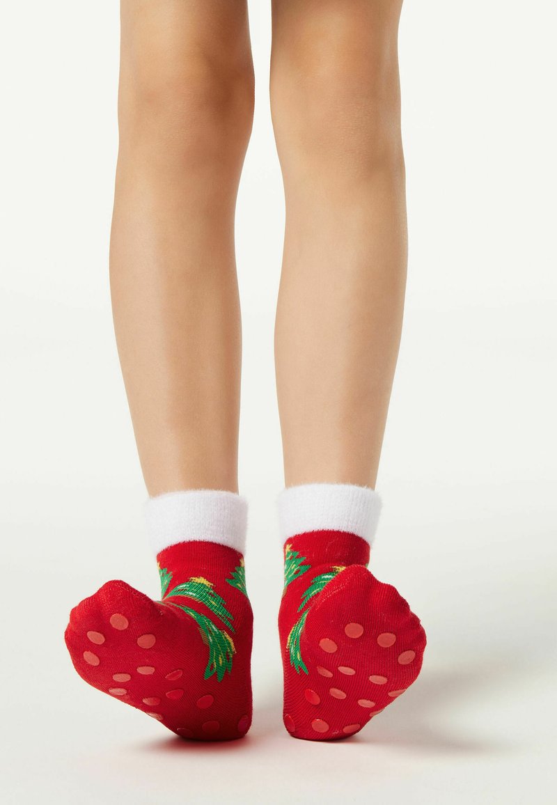 Červené ponožky s bílými manžetami, zdobené zelenými vzory listů a růžovými gumovými tečkami na podrážkách. Důraz na texturu a kontrast barev.