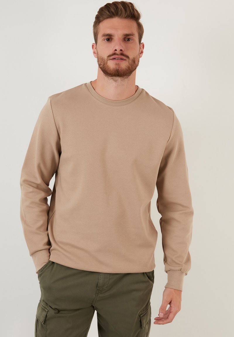 Sudadera de manga larga color marrón claro con cuello redondo y puños acanalados, presentada con pantalones cargo color verde oliva sobre un fondo neutro.