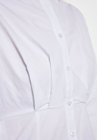 Weiße Bluse mit glatter Textur, ausgestattet mit einer Knopfleiste aus klaren Knöpfen und dezenten Fältchen in der Nähe der Brust.