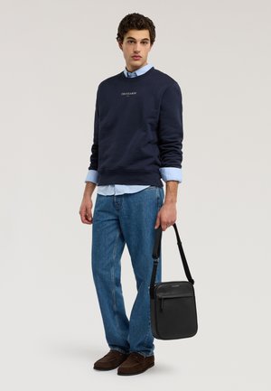 Ung mand iført navyblå sweatshirt over lyseblå skjorte og jeans, holder en sort læder skuldertaske mod en ensfarvet baggrund.