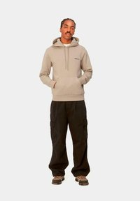Sudadera beige con bolsillo delantero y cordón, combinada con pantalones cargo negros holgados con bolsillos laterales. Usada con zapatos de diseño.