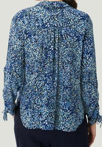 Blouse à manches longues, bleu marine, avec un motif floral agrémenté de touches de bleu clair et de vert. Tissu doux, encolure froncée et poignets de manches noués.