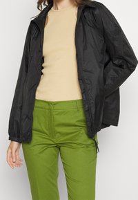 Czarna, lekka kurtka z zamkiem błyskawicznym z przodu i elastycznymi mankietami, zestawiona z beżowym, żeberkowanym topem na ramiączkach oraz zielonymi spodniami o dopasowanym kroju.