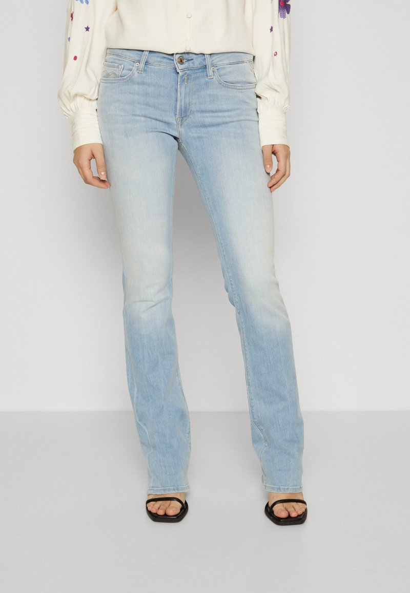 Femme portant un jean droit bleu clair, un chemisier crème avec broderies florales sur les manches, et des sandales noires à bout carré.
