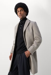 Abrigo de lana gris claro con solapas de muesca, cierre de un solo botón y bolsillos laterales, usado sobre un suéter de cuello alto negro y pantalones a rayas.