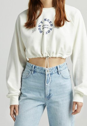 White cropped sweatshirt with navy text "DON'T NEED YOU I HAVE MEMES" and light blue high-waisted jeans worn by a woman.