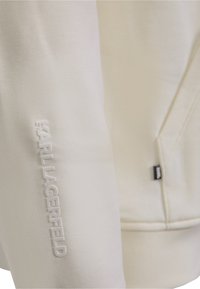 White sweatshirt featuring a textured fabric, embossed "KARL LAGERFELD" logo on the sleeve, and a small black tag near the seam.