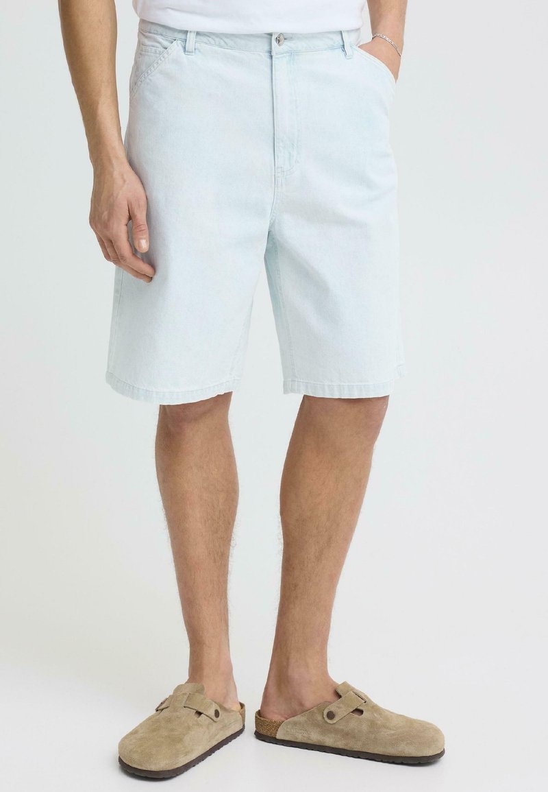 Homme portant un short en denim bleu clair et des chaussures beige en daim sans lacets, debout avec une main dans la poche contre un fond uni.