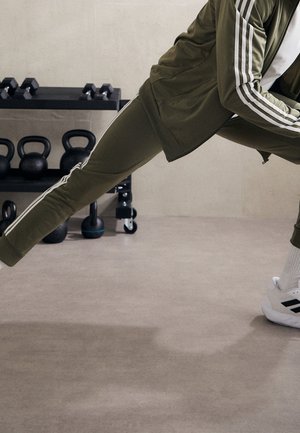 Osoba rozciągająca nogę do tyłu w zielonym dresie i białych trampkach przed kettlebellami i hantlami na stojaku w siłowni.