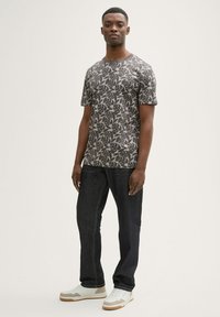 Szara koszulka z krótkim rękawem z ciemnym liściastym wzorem, w połączeniu z ciemnymi dżinsami i białymi trampkami z beżowymi akcentami oraz teksturowanymi podeszwami.