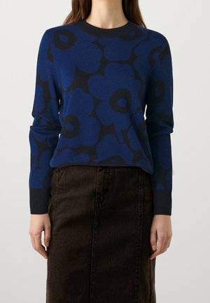 Kobieta ubrana w niebieski sweter z długim rękawem z dużymi czarnymi wzorami kwiatowymi oraz ciemnobrązową spódnicę z wysokim stanem.