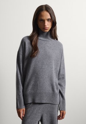 Mujer con cabello largo y castaño que lleva un suéter de cuello alto gris y pantalones grises a juego, de pie frente a un fondo claro y liso.