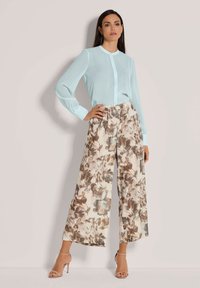 Lichtblauwe, doorzichtige blouse met knopen, gecombineerd met bloemen, wijd lopende broek in zachte beige, bruine en groene tinten; hoge hakken maken de look compleet.
