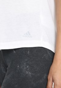 T-shirt bianco a maniche corte realizzato in tessuto leggero, con un logo Adidas grigio sottile sull'orlo. Abbinato a leggings scuri e testurizzati.