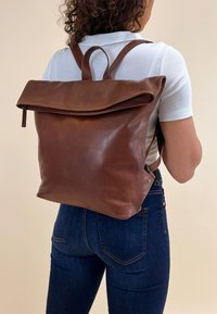 Brown leather backpack with a fold-over flap, zip closure, and two shoulder straps. Smooth texture, minimal design, and no visible hardware.