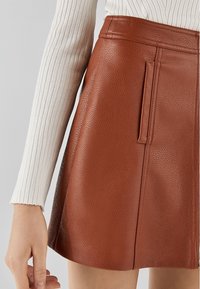 Hnědá kožená mini sukně s texturovaným povrchem, bočním kapsou a rovnou siluetou. V kombinaci s bílým žebrovaným dlouhým rukávem.