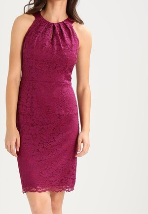 Femme portant une robe en dentelle magenta sans manches jusqu'aux genoux, avec un col froncé et un ourlet festonné, debout devant un fond uni.
