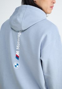 Světle modrá mikina s kapucí vyrobená z měkké látky, s velkou kapucí, bílým textem "Motorsport" a logem BMW na zadní straně.