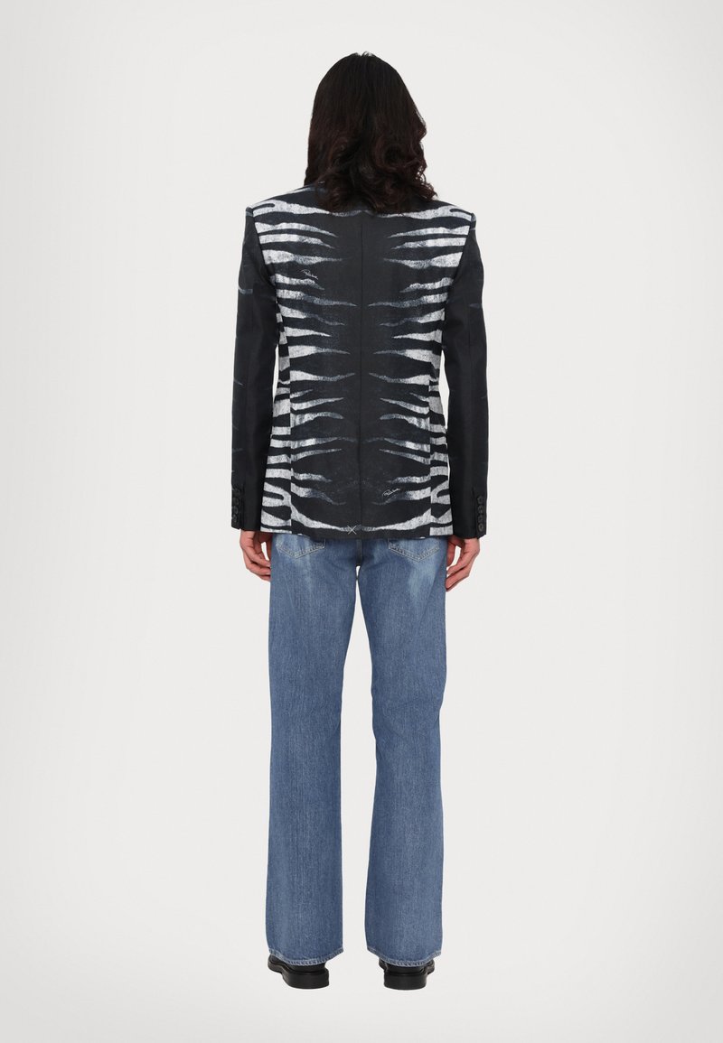 Sort blazer med et hvidt abstrakt mønster på ryggen, parret med blå flare jeans. Materialet virker glat med struktureret skrædderarbejde.