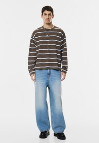 Jonge man in een bruin en wit gestreept longsleeve shirt, lichtblauwe wijde jeans, zwarte schoenen en een zilveren ketting staande tegen een witte achtergrond.