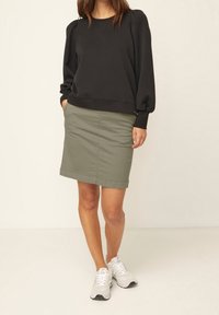 Femme portant un top noir à manches longues, une jupe olive jusqu'aux genoux et des baskets blanches, se tenant avec les mains dans les poches sur un fond blanc.