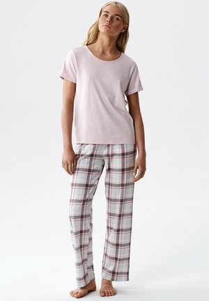 Młoda kobieta w jasnoróżowym, krótkim topie i pastelowych, kraciastych piżamowych spodniach, stojąca boso na jednolitym tle.