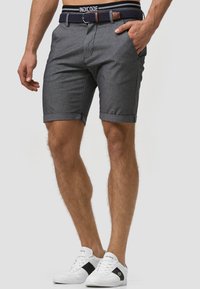 Shorts en tissu gris tissés avec une coupe ajustée, des revers roulés, des poches frontales et une ceinture tressée bleu marine avec des accents marron. Portés avec des baskets blanches.