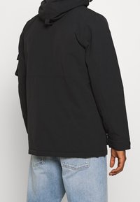 Černá kapucová bunda s hladkým, matným povrchem, která má uvolněný střih, zipové zapínání a boční kapsy. Z pohledu zezadu nejsou viditelné žádné značky.
