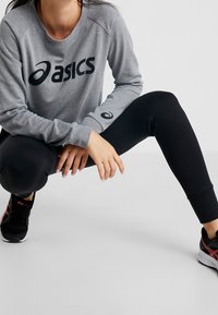 Felpa grigia Asics con logo nero, leggings neri e scarpe da ginnastica nere con dettagli rossi. Modella in posizione accovacciata.