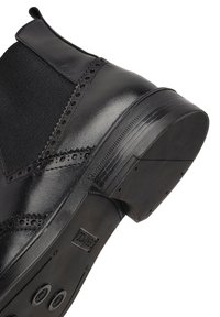 Bota negra de cuero hasta el tobillo con paneles elásticos en los laterales, detalles decorativos de brogue y puntera redondeada. Suela de goma texturizada con ranuras.