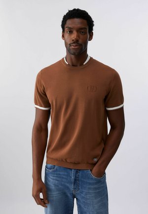 Hombre con cabello corto y rizado que lleva un suéter marrón de manga corta con ribete blanco y jeans azules, de pie con una mano en el bolsillo.