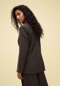 Blazer marrone scuro a righe con una vestibilità su misura, spalle strutturate e chiusura a un bottone, mostrato su uno sfondo neutro.