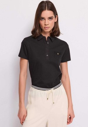 Giovane donna con capelli castani fino alle spalle che indossa una polo nera a maniche corte e pantaloni bianchi con coulisse su sfondo bianco.