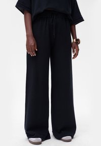 Zwarte, wijde broek gemaakt van textuurstof, met een elastische tailleband en een trekkoord. Gecombineerd met witte sneakers.