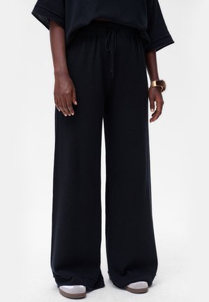 Pantalons larges noirs en tissu texturé, avec une taille élastique et un cordon de serrage. Associés à des baskets blanches.