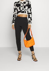 Zwart-witte graphic crop trui met lange mouwen, gecombineerd met strakke zwarte broek en een glanzende oranje gewatteerde handtas. Zwarte sandalen met bandjes.