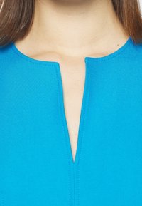 Blaues Kleidungsstück mit einem runden Halsauschnitt und einem Schlüssellochöffnungsdesign, gefertigt aus einem glatten Stoff und mit sauberen Nähdetails.