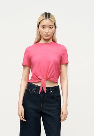 Jeune femme aux longs cheveux blonds portant un haut court rose noué et un jean taille haute bleu foncé, debout sur un fond blanc.