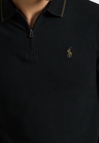Polo noir zippé avec un col court et un petit logo brodé représentant une personne montant à cheval sur la poitrine.
