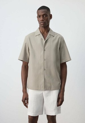 Homme portant une chemise beige à manches courtes boutonnée et un short blanc jusqu'aux genoux, debout devant un fond clair uni.