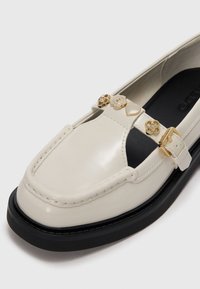 Chaussure en cuir verni crème à bout rond, ornée d'accents floraux dorés décoratifs et d'une semelle plateforme noire.